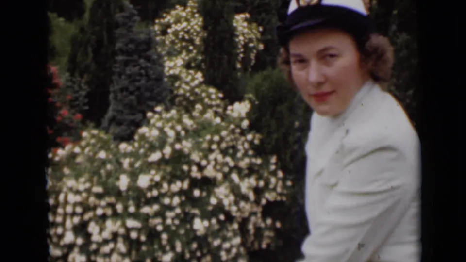 A woman in 1940s dress and hat gives a small smile toward the camera beside a lush flower bush in archival 8mm footage.