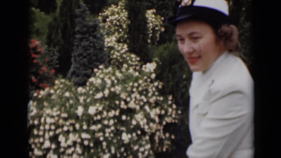 The woman looks down with a soft smile in the garden, her white jacket and hat contrasting with the dark trees and bright blossoms.