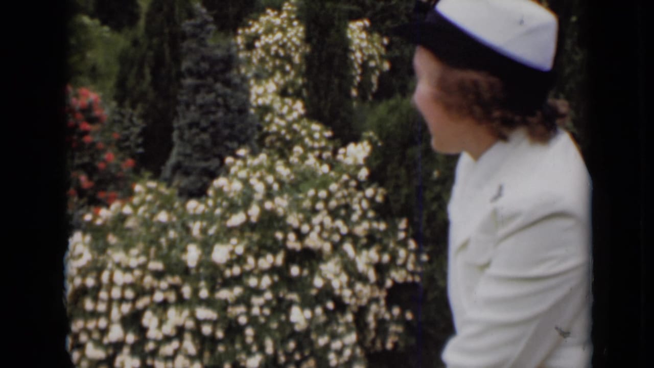 Woman in a white uniform and cap turns beside flowering garden shrubs.