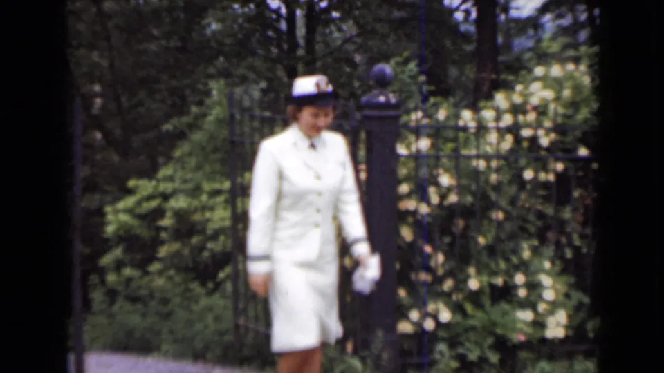 The woman walks toward the camera in a white uniform and gloves, framed by an iron gate and flowering plants.