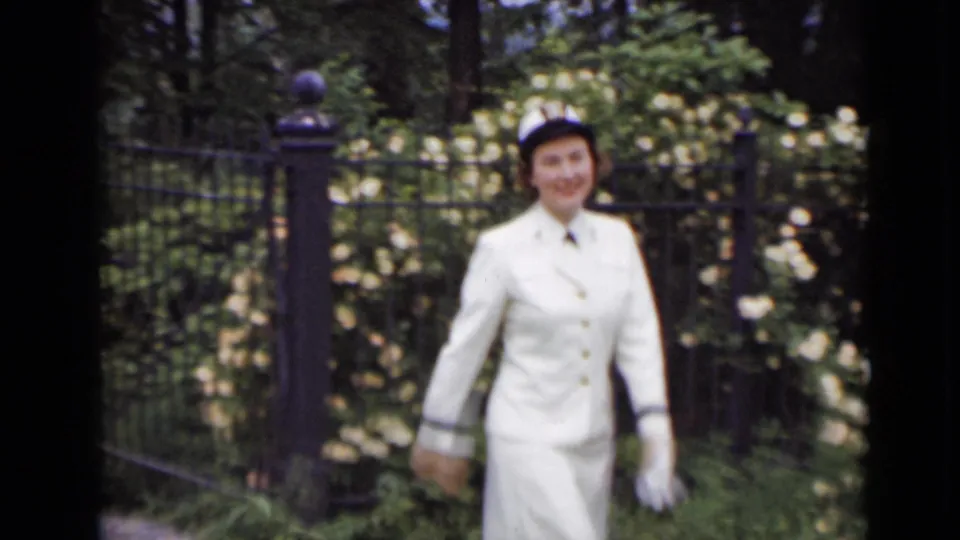 A smiling woman in a white uniform and cap approaches the camera near a black iron gate in the garden.