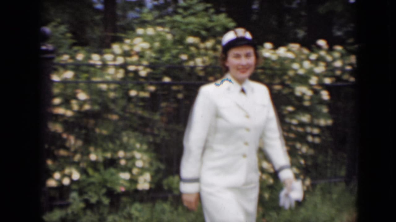 Woman in a white uniform and cap standing near a garden fence and white flowers.