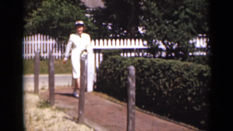In 1947 New York footage, the woman in a white uniform walks along a garden path past posts, hedges, and a white picket fence.