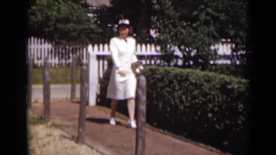 The woman in a white uniform and cap stands on a garden path in 1947 New York footage, framed by hedges and a white picket fence.