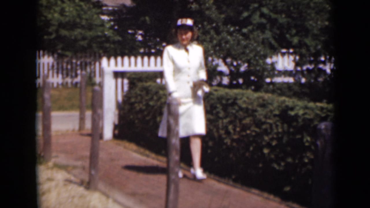 Woman in white outfit and hat stands on a brick path beside hedges and a white fence.
