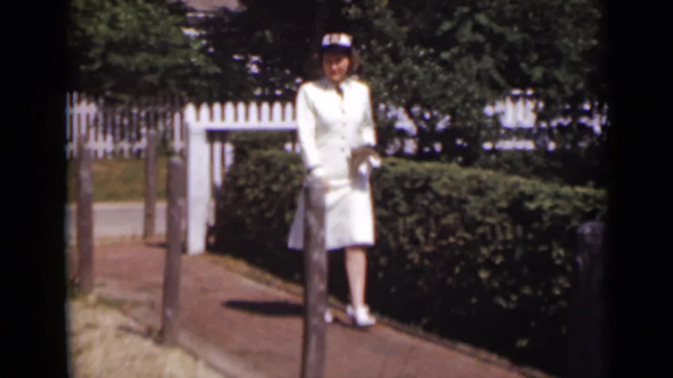 In 1947 New York home-movie footage, a woman in a white outfit and matching hat stands on a sunny brick path near hedges and a picket fence.