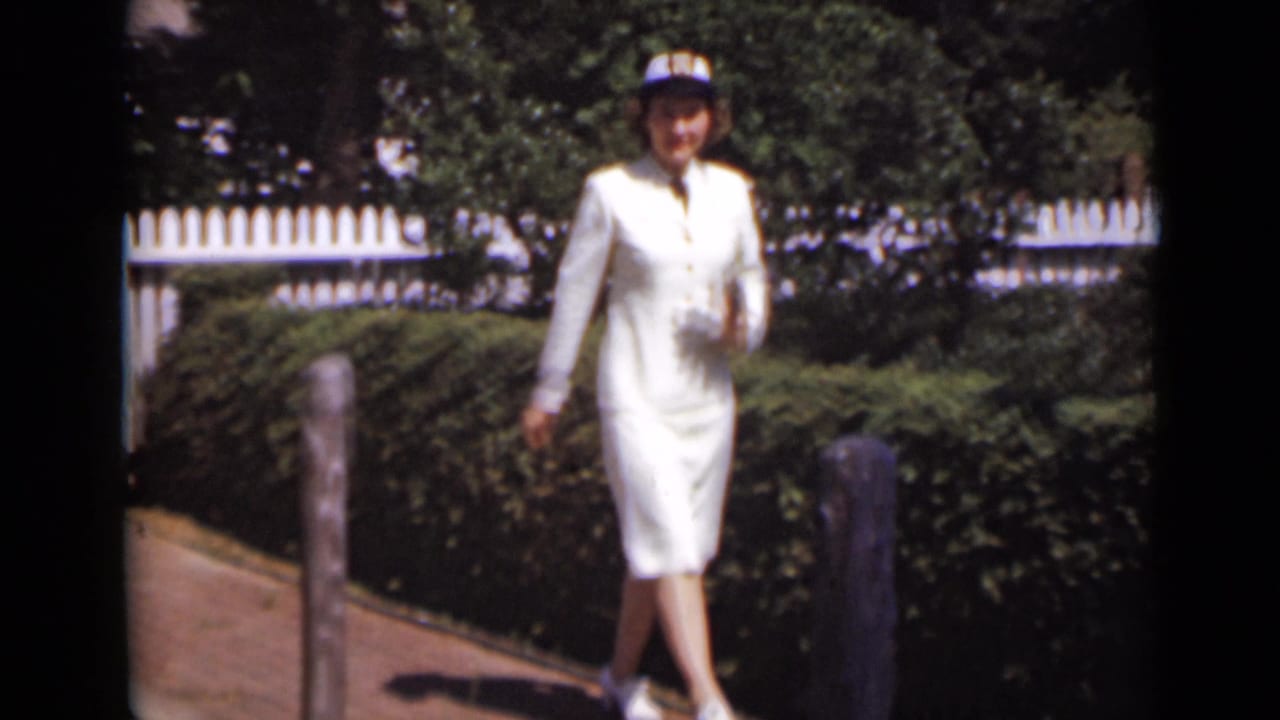 A woman in a white outfit and hat walks along a sunny garden path in New York, with hedges, posts and a white fence behind her.
