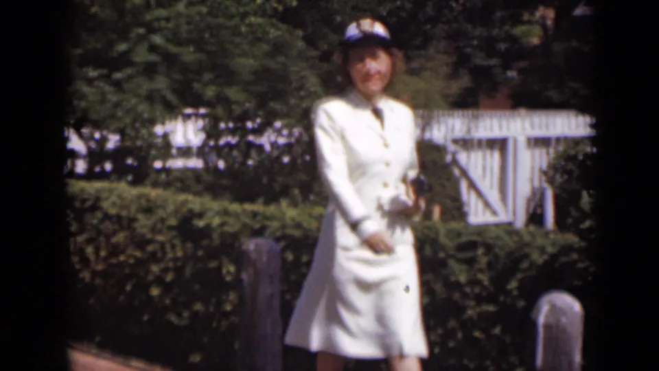 The woman in white moves closer to the camera on the brick path, framed by hedges, wooden posts and a white garden gate.