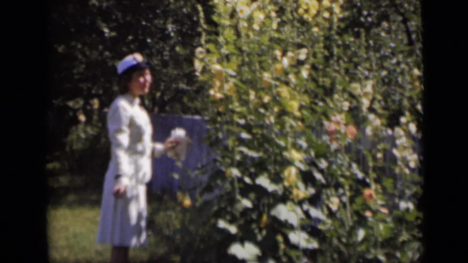 A woman in a white outfit and hat stands near tall flowering plants and dense garden greenery in the 1947 New York footage.