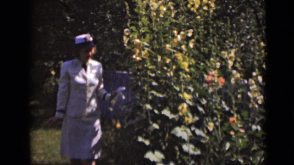 A softly focused 8mm frame shows the woman closer to the plants, with her hat and light clothing visible against the garden foliage.