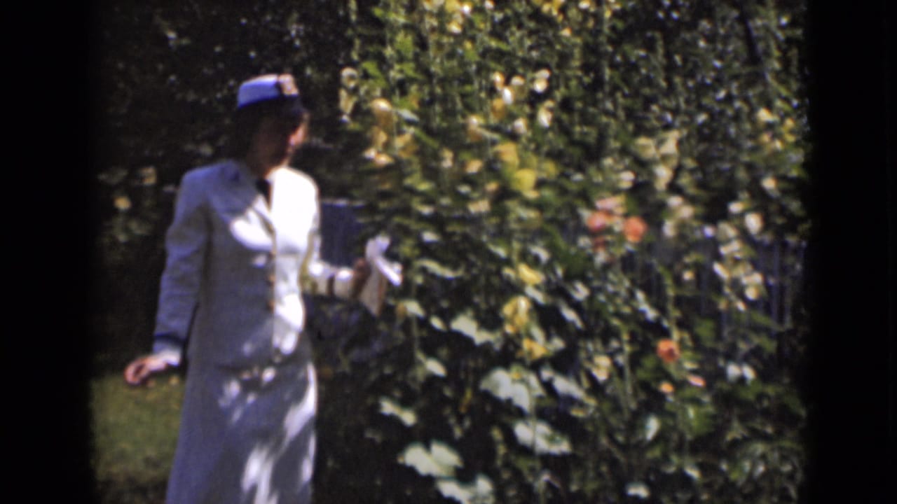 A woman in 1947 New York home-movie footage walks through a sunlit garden in a pale suit and brimmed hat.