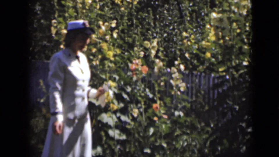 The woman pauses or turns beside dense garden plants, holding a small pale object in her hand.