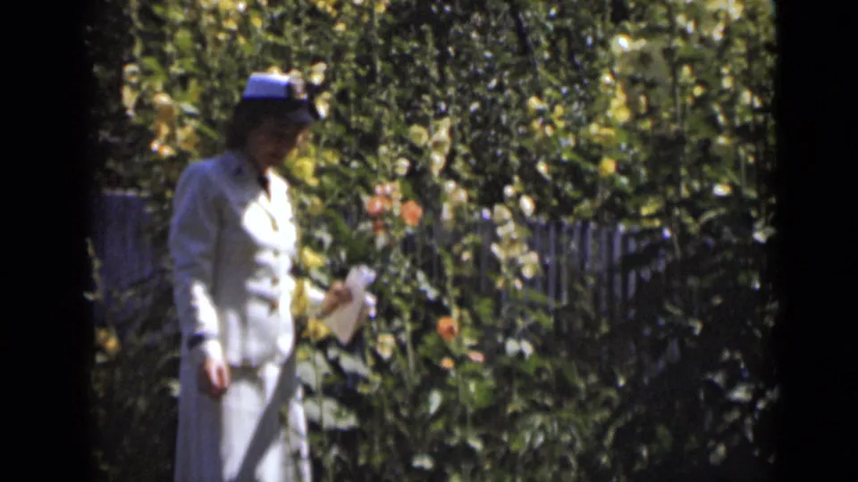 A profile view shows the woman moving along a garden path or border with flowers and foliage behind her.
