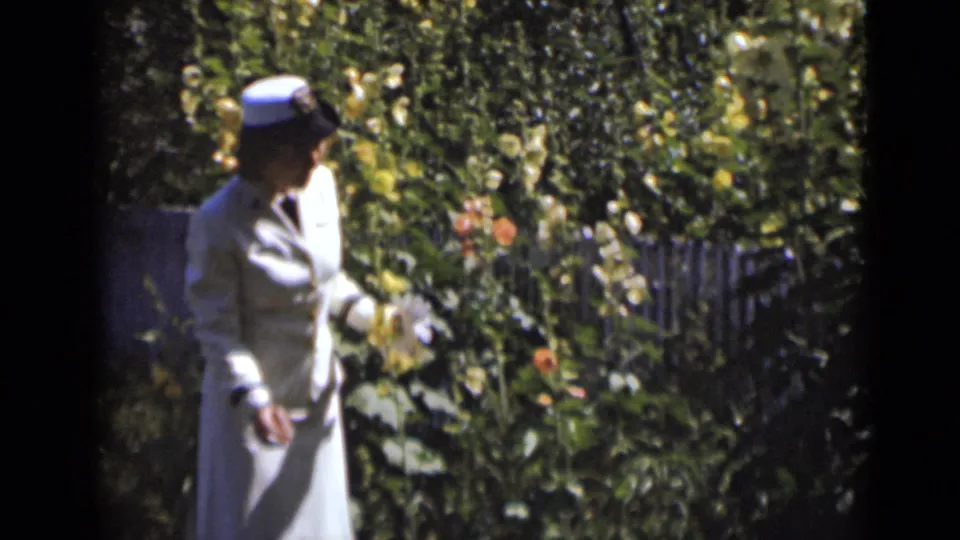 The woman continues her slow garden walk beside dense greenery and scattered orange flowers in bright sun.