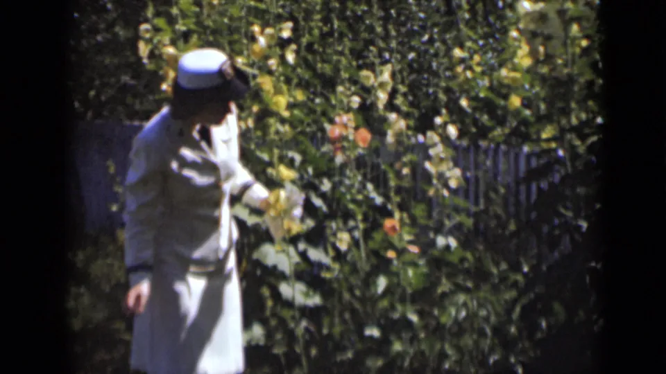 Adult woman in 1947 New York home-movie footage stands in a sunlit garden wearing a light dress and hat.