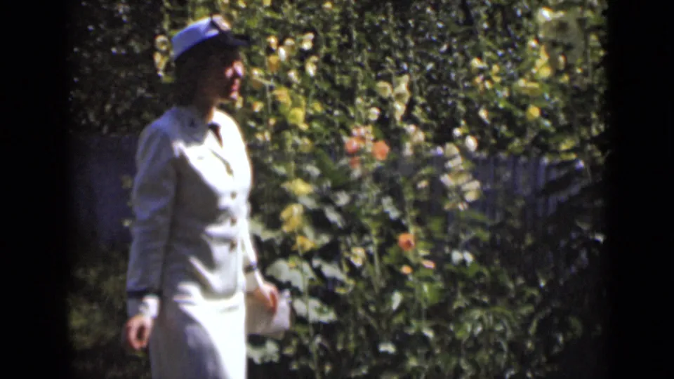 Adult woman in light mid-century attire stands near flowers and greenery in 1947 New York home-movie footage.