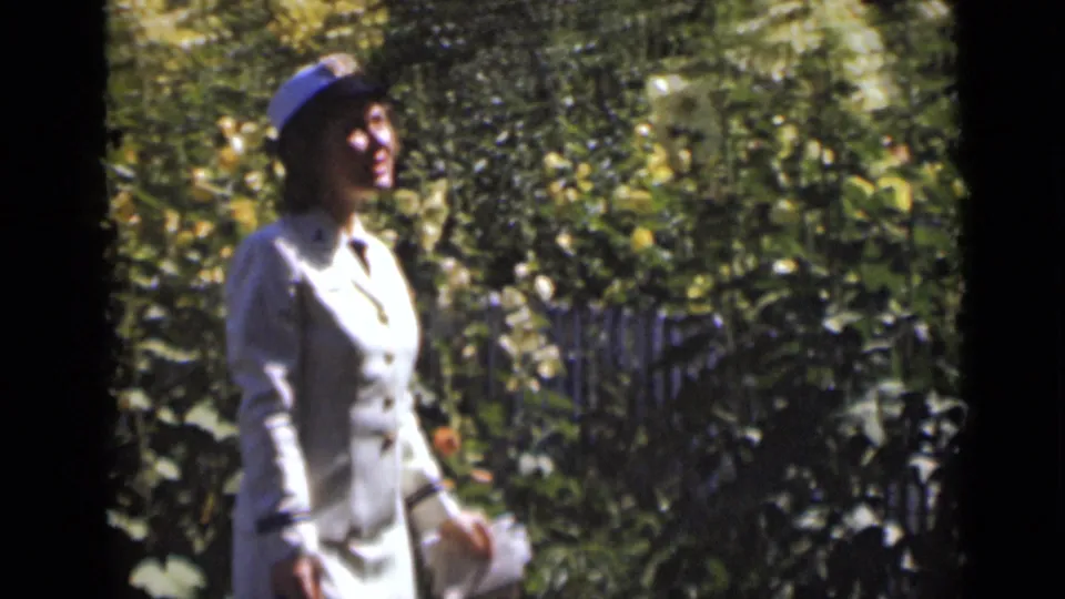 Adult woman in a light dress and hat walks through a sunlit New York garden in 1947 home-movie footage.