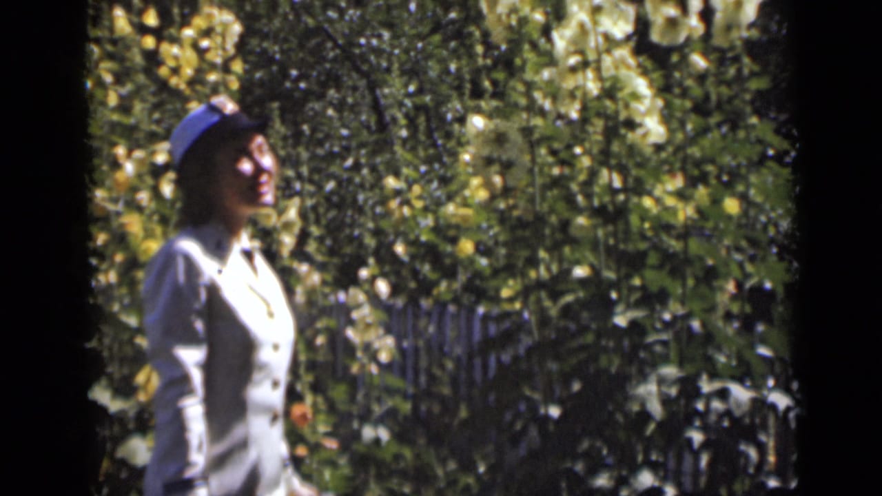 Woman in a light uniform and cap walking beside tall flowering plants.