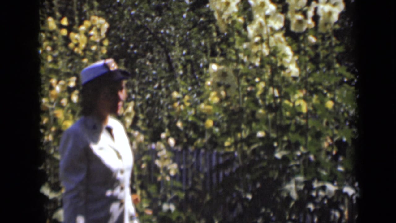 Uniformed woman in profile walking past flowers and garden foliage.