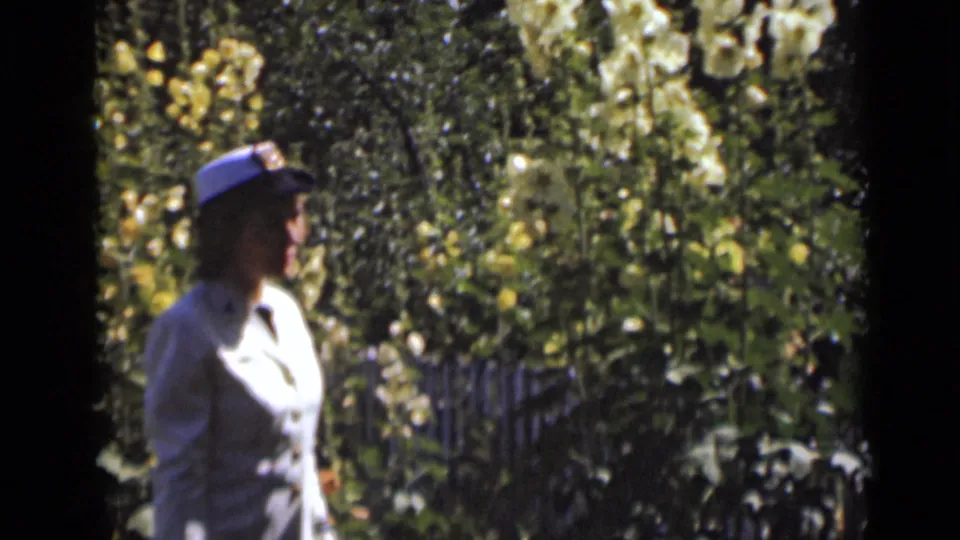 The woman appears in profile as she moves past dense garden foliage and pale flowers in New York archival home-movie footage.