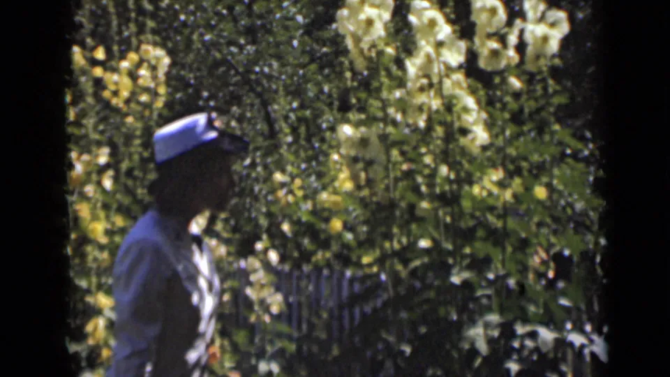 The uniformed woman is seen partly in shadow near tall flowers and dense greenery in the garden sequence.