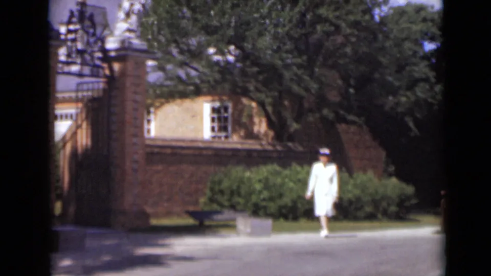 The woman walks across paved grounds near a brick building, shrubs, trees, and an ornate gate in the New York 1947 clip.