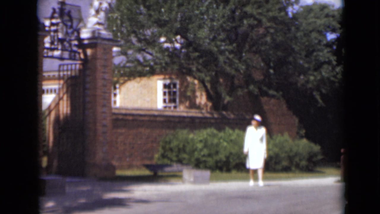 In 1947 New York, a woman in a white dress and hat stands near a brick gate, shrubs, trees, and a paved entrance path.
