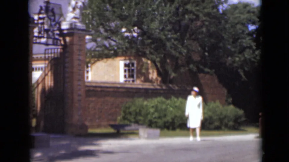 In 1947 New York, a woman in a white dress and hat stands near a brick gate, shrubs, trees, and a paved entrance path.
