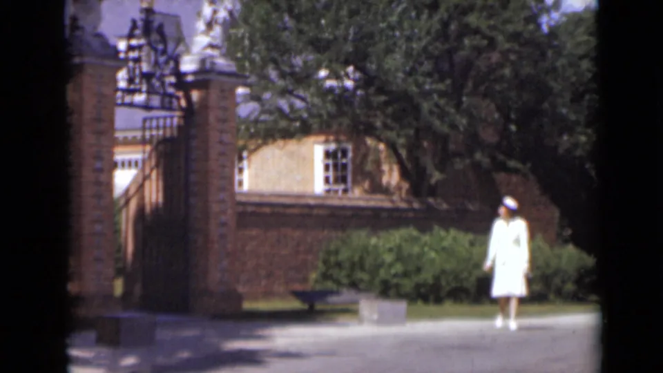 The woman remains near the entrance path in a 1947 New York exterior, framed by a brick wall, shrubs, trees, and gate masonry.