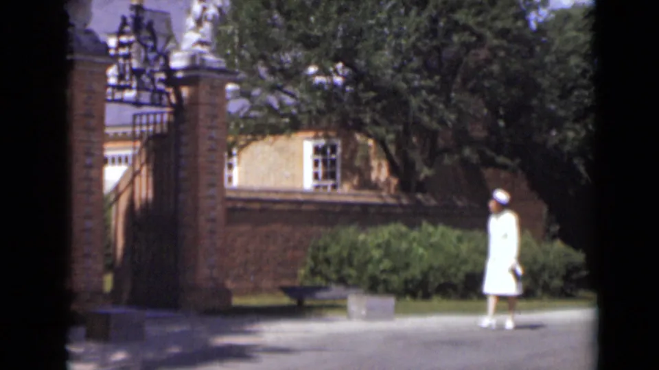 A woman in white walks along the paved path near a brick gate and leafy grounds in this 1947 New York home-movie frame.