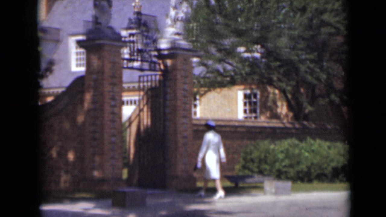 Woman in white nears the gate beside brick pillars and greenery.