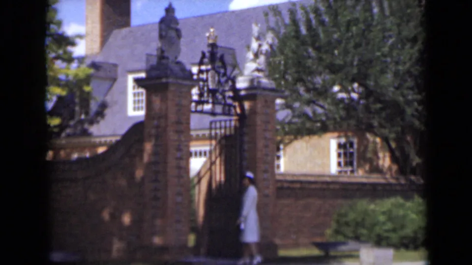The woman reaches the gate area, partly framed by the brick wall, ironwork, trees, and sunlit facade in the background.