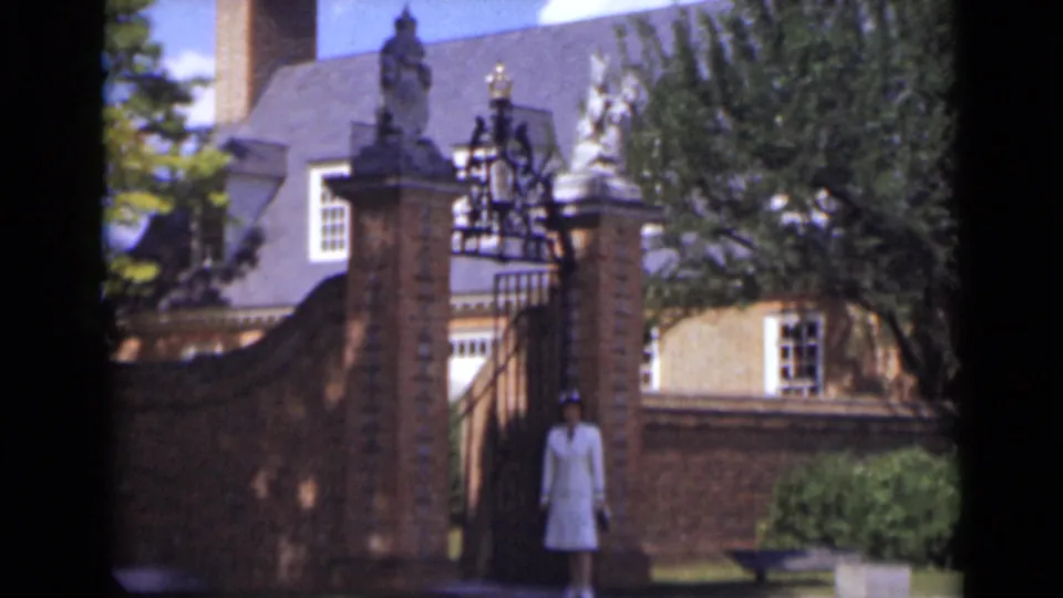 The woman in white stands near the ornate brick-and-iron gate, facing the camera area with shrubs and a formal facade behind her.