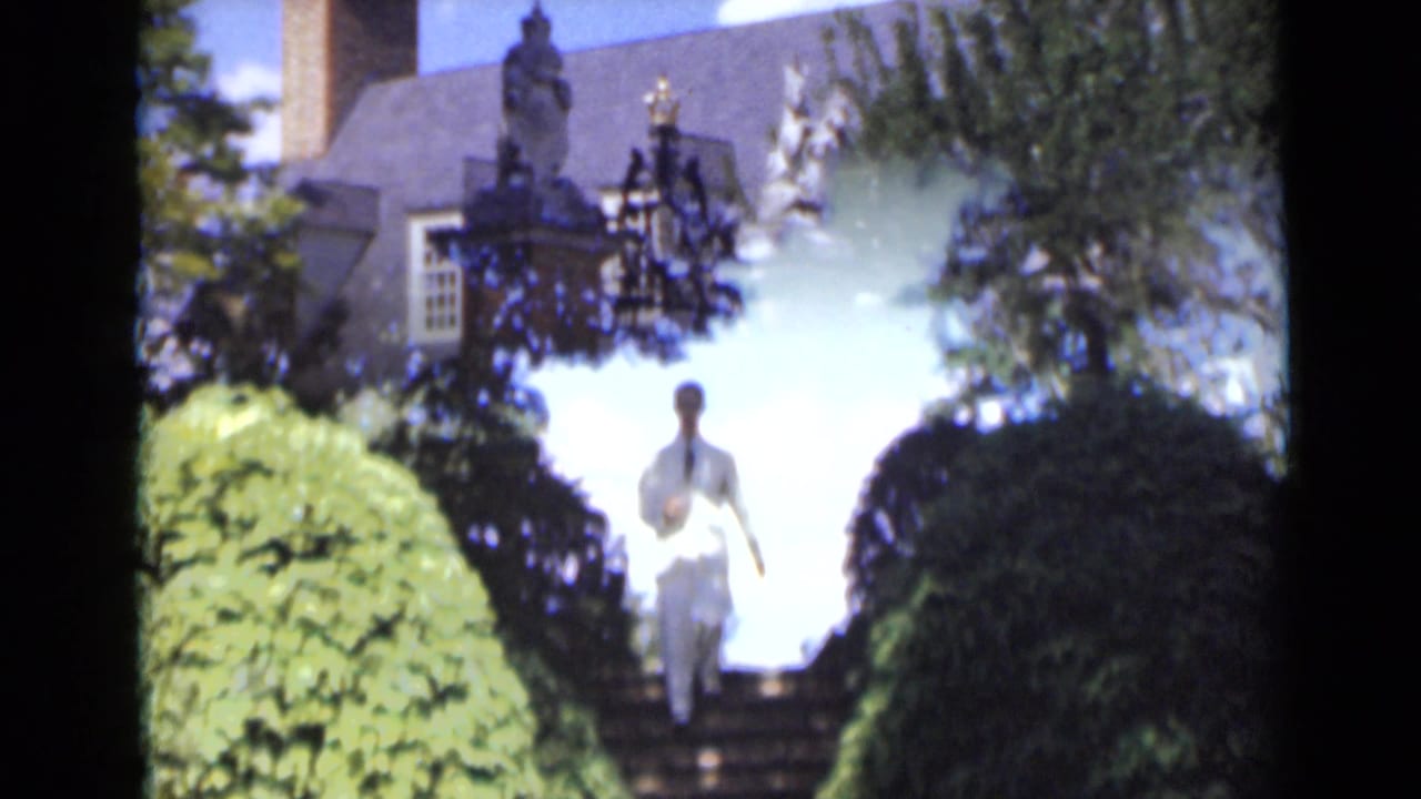 Man in a white suit descends garden steps between tall hedges near a large house.