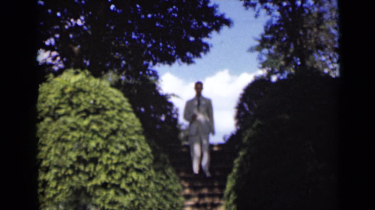 The man continues down the garden staircase in a white suit, with dense hedges, tree shade, blue sky, and clouds around him.