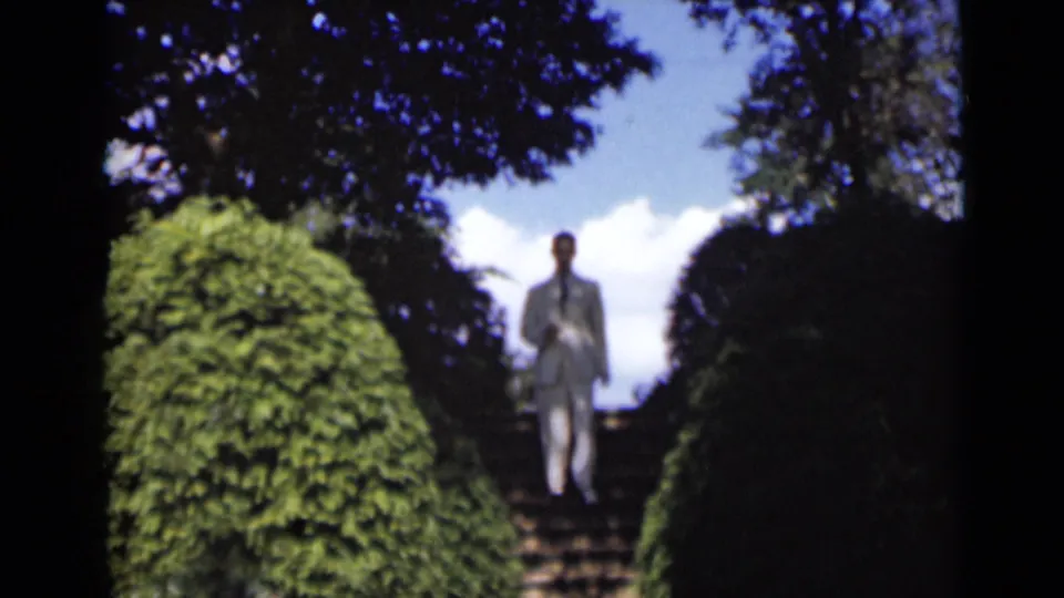 The man continues down the garden staircase in a white suit, with dense hedges, tree shade, blue sky, and clouds around him.