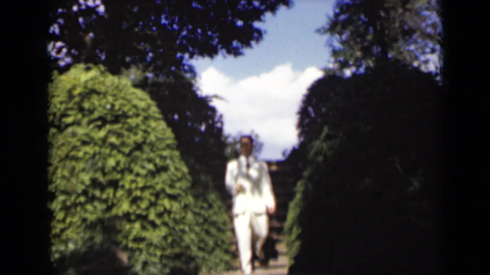 Near the end of the shot, the man in a white suit and hat reaches the lower part of the stone staircase between tall green hedges.