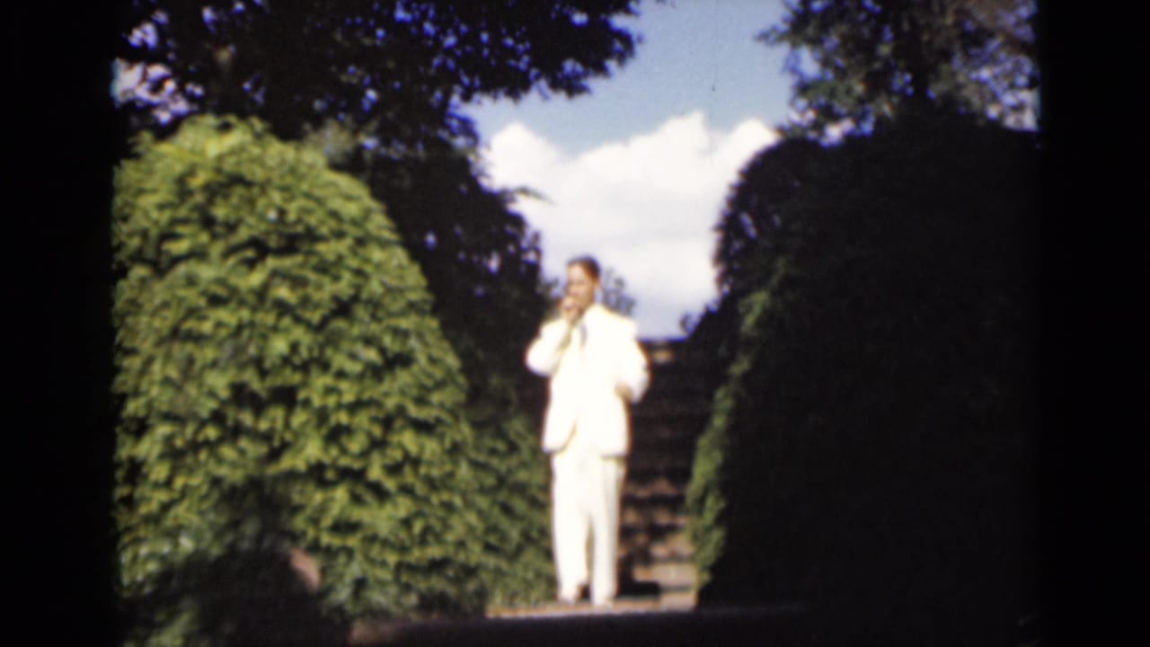 Man in a white suit stands near the top of garden stairs between hedges.