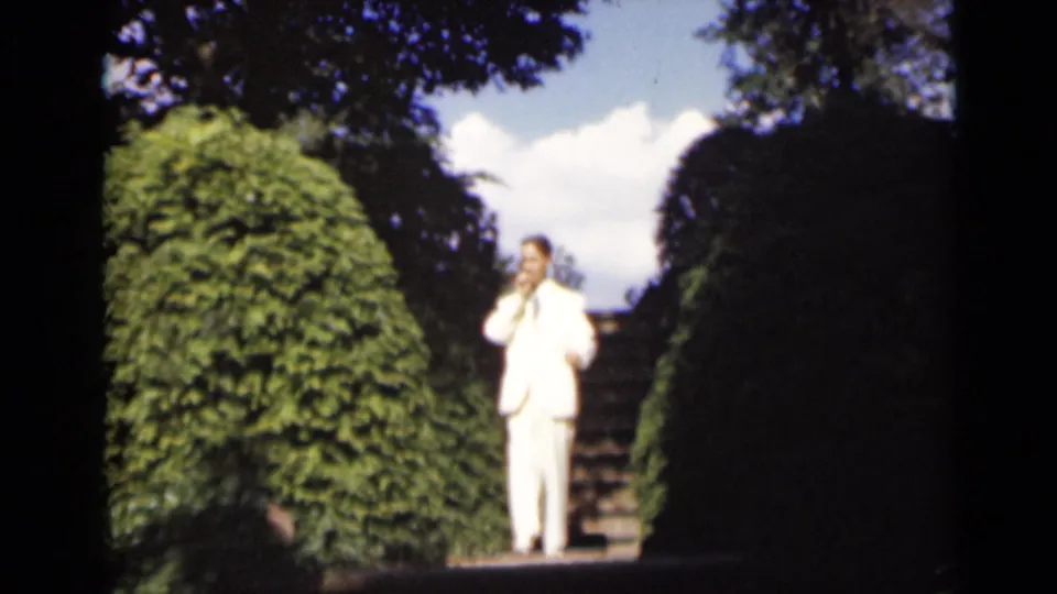In a 1947 New York 8mm frame, a man in a white suit pauses near stone garden stairs flanked by dense hedges.