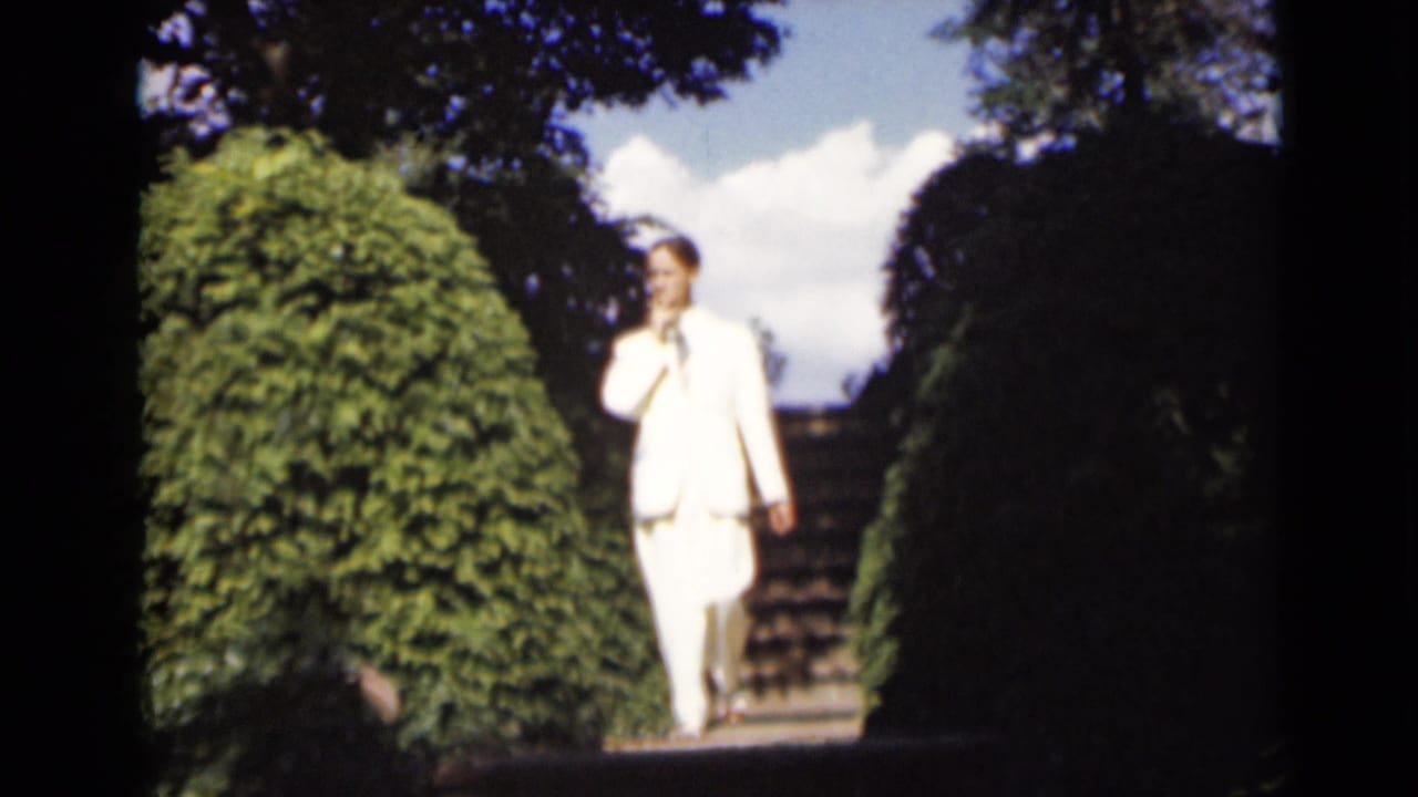 The man descends the garden steps in a white suit, with hedges and bright sky visible in the 1947 New York home-movie frame.