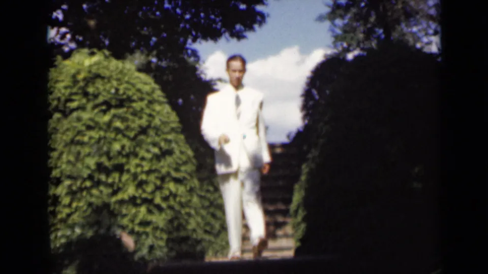 A closer 8mm view shows the suited man walking down stone garden stairs, framed by clipped hedges and overhead shade.