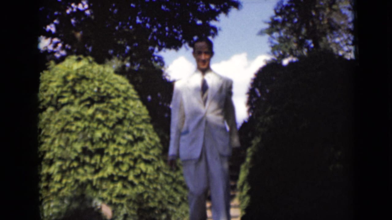 Man in a white suit standing on outdoor steps between tall green shrubs.