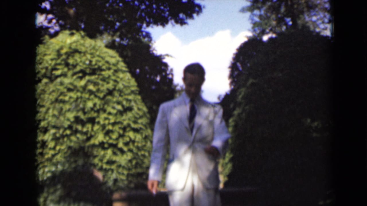 Man in a white suit walking down steps beside rounded green shrubs.