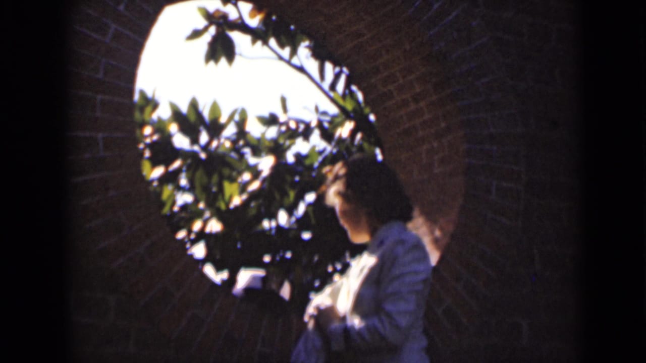 In 1947 New York home-movie footage, a woman in a light shirt stands in profile near a round brick opening with sunlit leaves beyond.