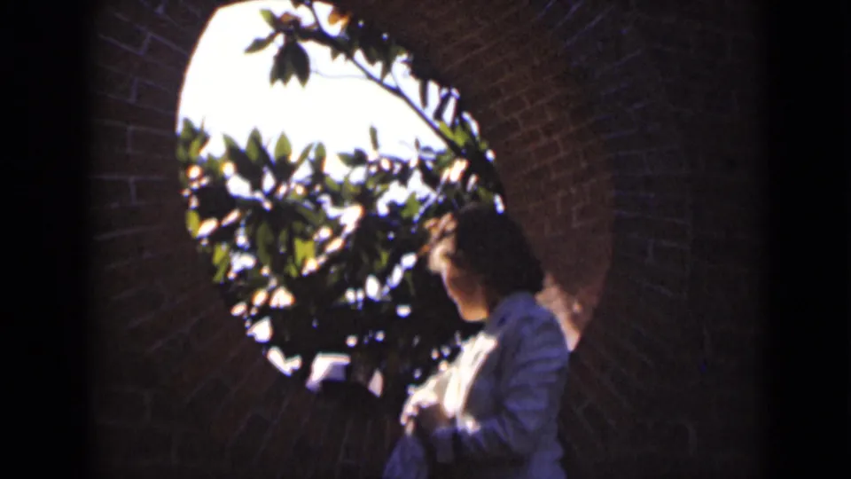 In 1947 New York home-movie footage, a woman in a light shirt stands in profile near a round brick opening with sunlit leaves beyond.