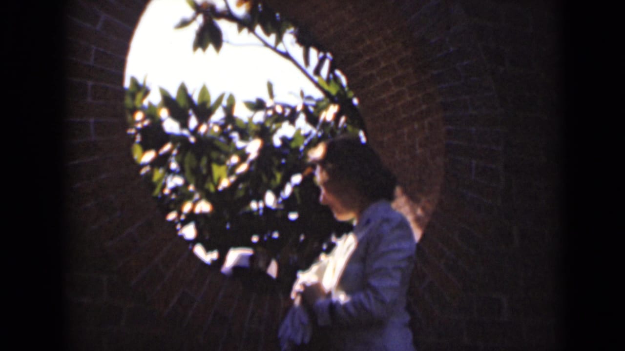 Woman looks toward a bright round opening framed by brick and leafy branches.