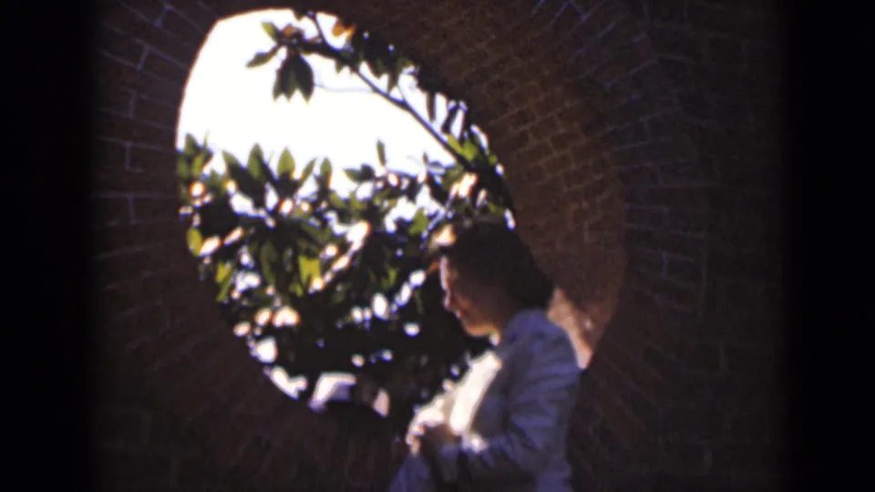 A softly focused 8mm frame shows the woman beside the circular brick opening, with dark leaves silhouetted against the sunlit exterior.
