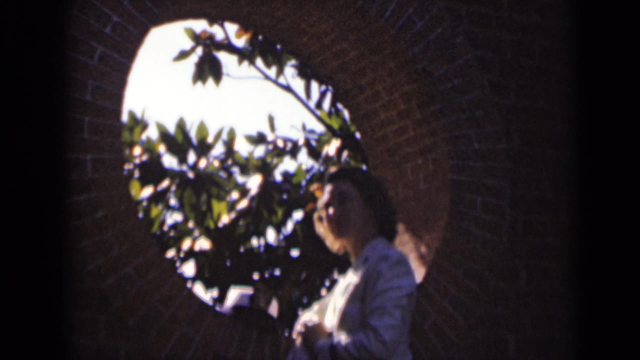 Woman in a white uniform seen through a brick archway with leaves and bright sky behind her.