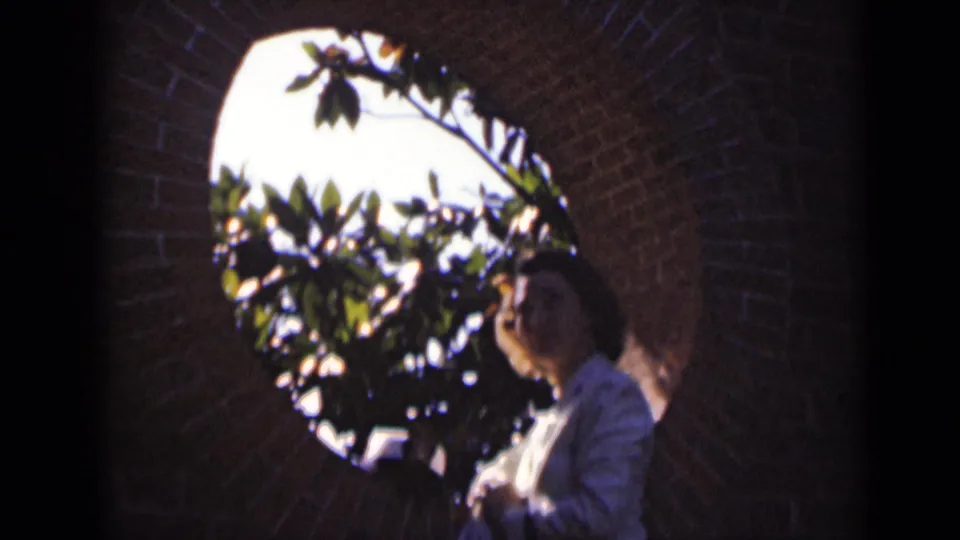 A woman in a white nurse-style uniform stands under a brick archway, framed by leaves and bright outdoor light in New York home-movie footage.