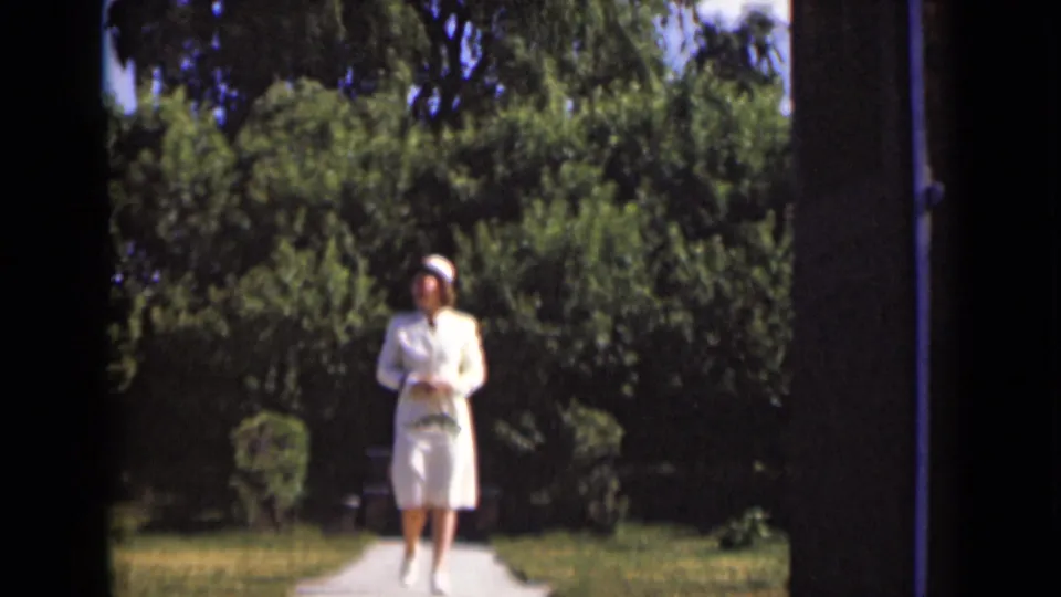 1947 New York frame showing a woman in a white nurse-style uniform walking along a garden path bordered by shrubs and trees.
